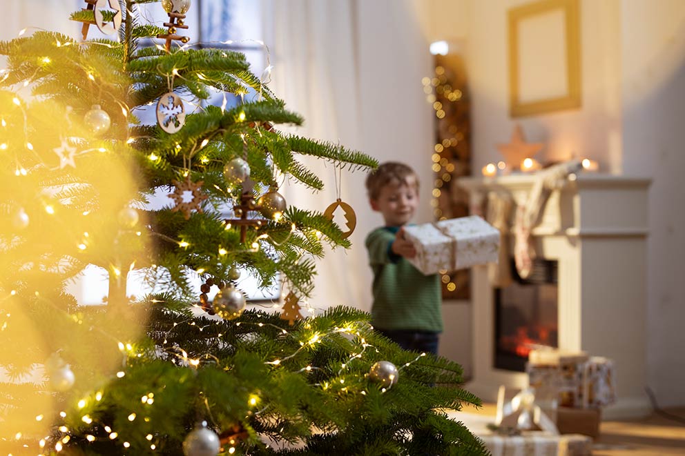Een jongetje draagt een cadeautje naar de versierde kerstboom.