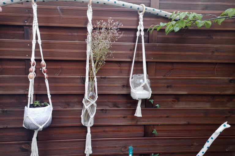 Drie geknoopte plantenhangers hangen aan een stok voor een schutting; in de rechter plantenhanger is een theelichtje aangebracht.