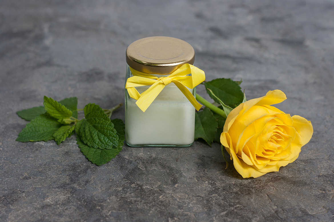 Crème deodorant in een glazen potje met een geel strikje, ernaast een gele roos en citroenmelisseblaadjes