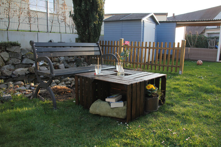 De tuintafel van houten kisten staat op het gazon voor een tuinbank.