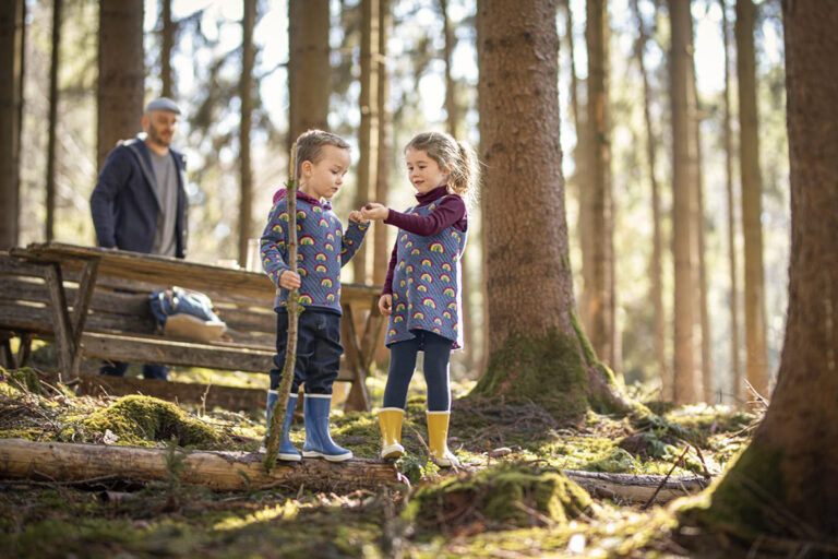 Twee kinderen verkennen samen het bos en zijn bosdieren, op de achtergrond is hun vader te zien.