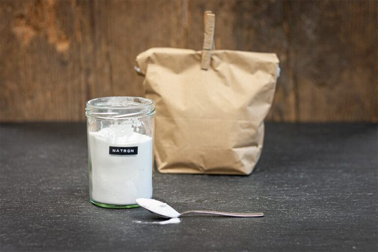 Een lepel natron/baking soda ligt naast een open glas met op de achtergrond een papieren zak