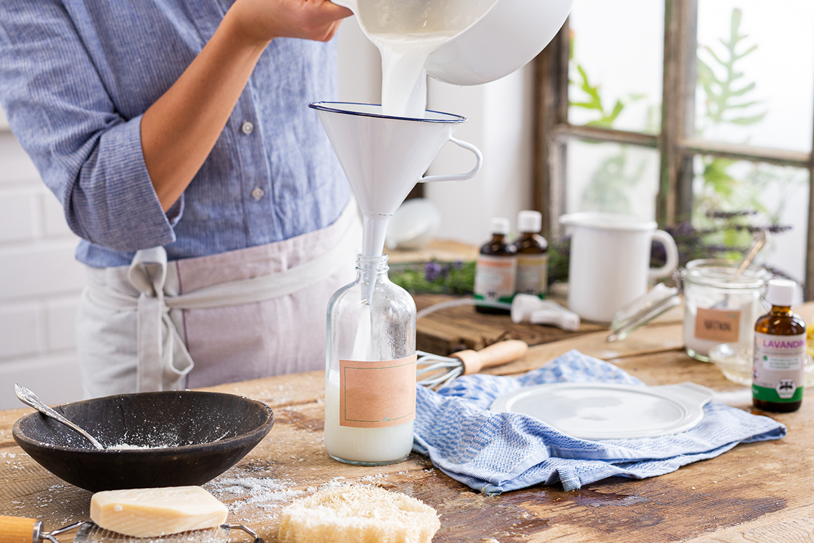 Een vrouw staat in een keuken en giet zelfgemaakt afwasmiddel door een trechter in een glazen fles.