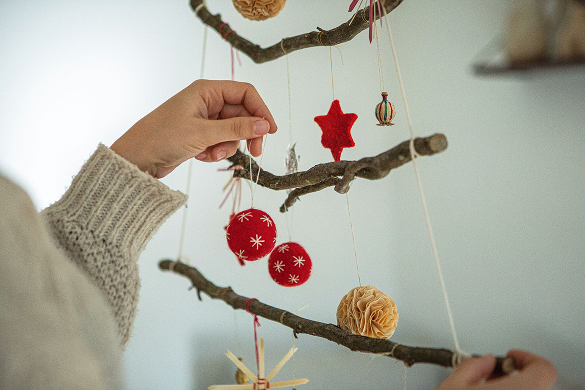 Boomversieringen hangen al aan de kerstboom van takken, een hand hangt net een rode vilten bal aan een tak.
