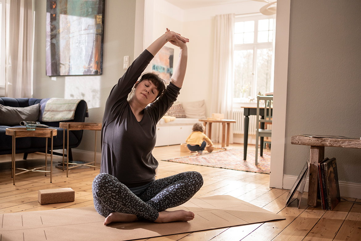 Een vrouw zit op een kurken yogamat in de woonkamer en strekt haar armen zijwaarts boven haar hoofd.