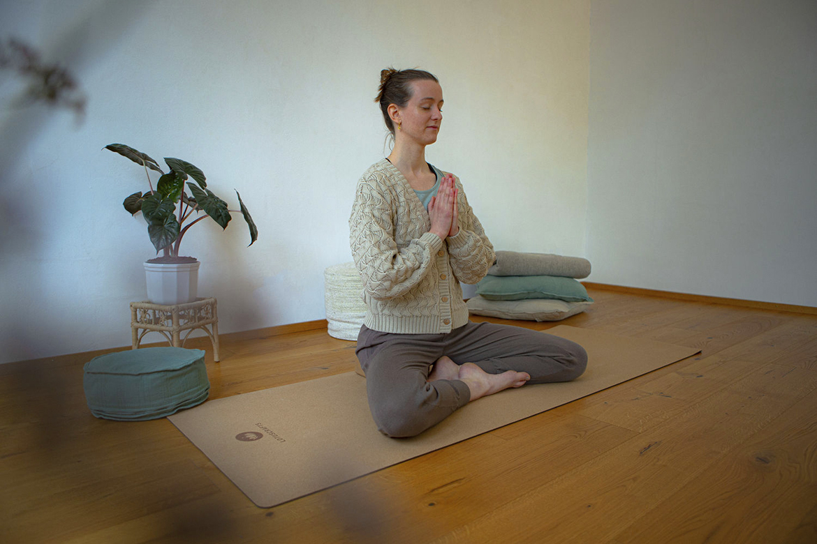 Een vrouw zit in kleermakerszit op een kurken yogamat, met haar handen voor haar borst gevouwen.