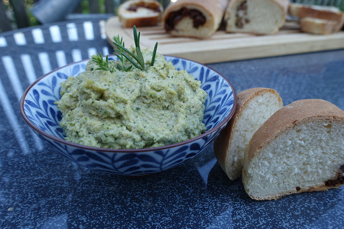 Een kom met courgettespread staat naast zelfgemaakte ciabatta op de blauwe tafel.