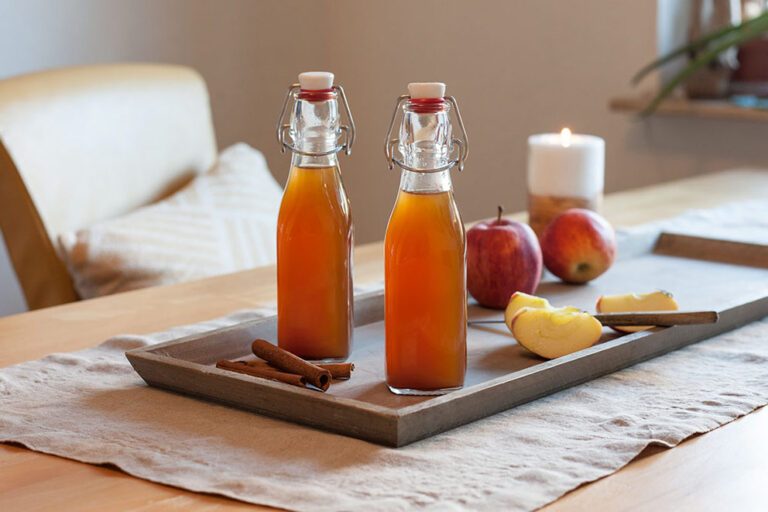 Twee flessen gevuld met zelfgemaakte appel-kaneellikeur staan op een houten schaal op een gedecoreerde tafel.