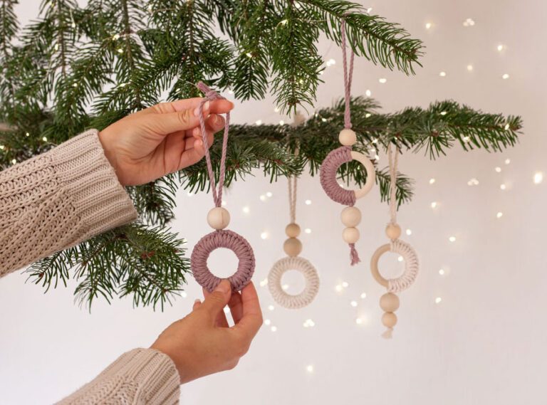 De macramé kerstboomversiering wordt met één hand opgehangen aan een dennentak.