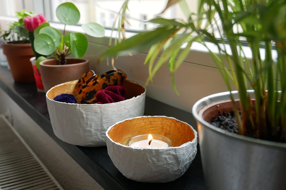 Zelfgemaakte schalen van papier-maché staan op een vensterbank tussen enkele planten.
