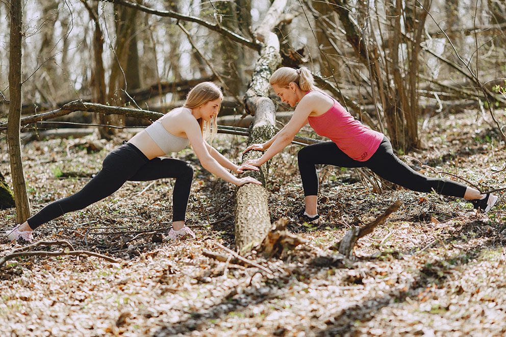 Twee vrouwen leunen op een boomstam om zich te strekken bij het sporten in het bos.