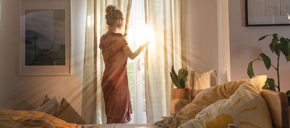 Een jonge vrouw staat 's morgens vroeg voor het raam en de eerste zonnestralen vallen op haar gezicht.