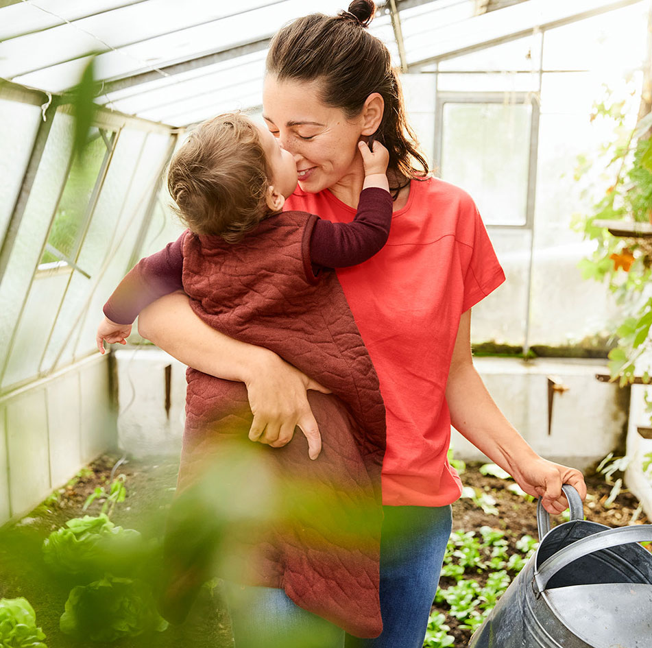 Baby in de gewatteerde slaapzak op de arm van mama in een plantenkas