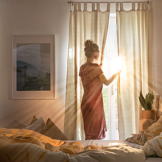 Een jonge vrouw staat 's morgens vroeg voor het raam en de eerste zonnestralen vallen op haar gezicht.