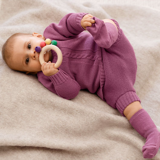 Baby in een rozenhout kleurige scheerwollen kledingset van Waschbär speelt met een houten bijtring.