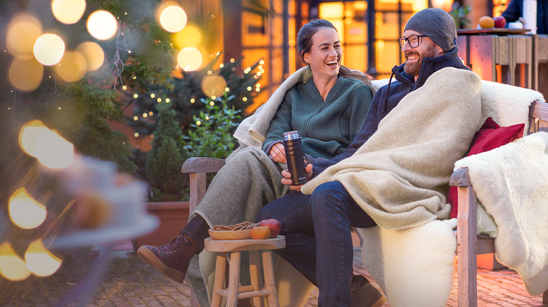 Een man en vrouw zitten ingewikkeld in een plaid buiten op een bank en genieten van de kerstsfeer.